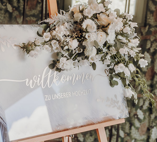 Auf einem mit Blumen dekorierten Schild steht "Herzlich willkommen zu unserer Hochzeit"